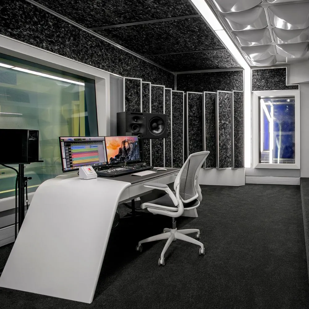 A recording studio with a white curved desk, black acoustic walls and a window into the booth
